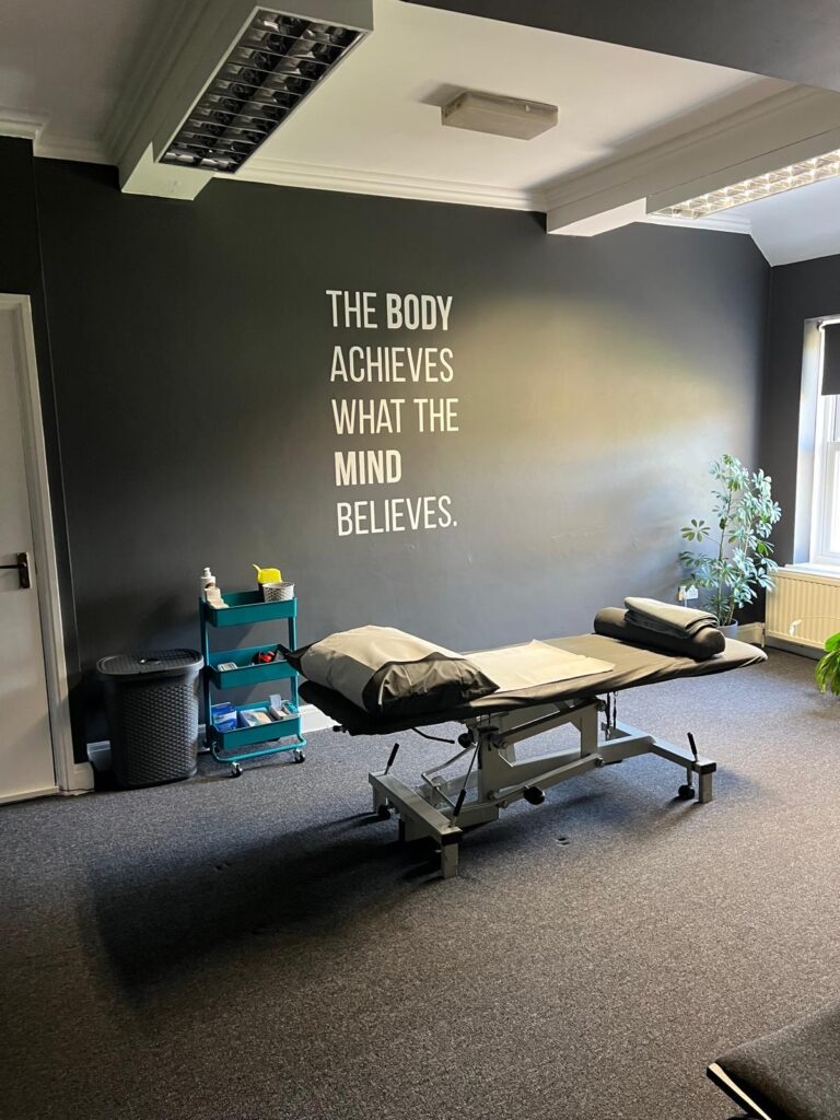 A therapy room featuring an adjustable treatment bed, a motivational quote on the wall, and a plant near the window.