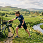 A cyclist stretches beside a green bike in a lush countryside scene, with rolling hills and a flowing river adorned with daffodils.
